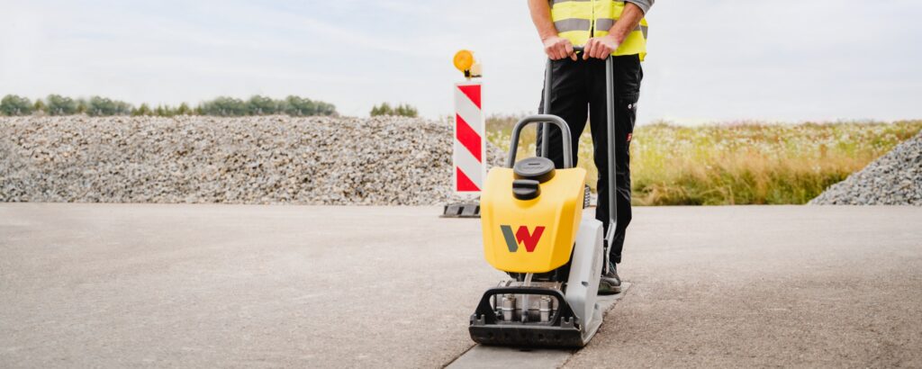 Operator compacting asphalt with a Wacker Neuson BPS forward plate beside a road barrier
